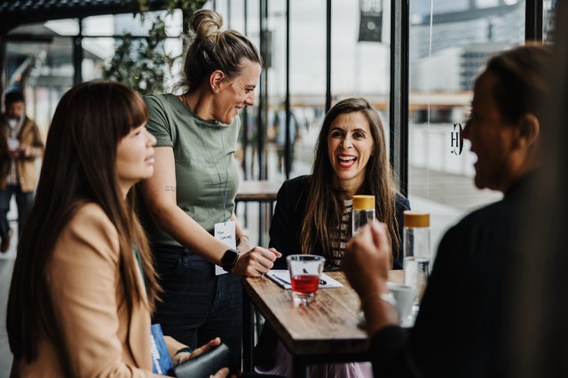 Women engaging in lively discussion at DL24–M, seated at a wooden table in a modern cafe setting, with natural light streaming through large.